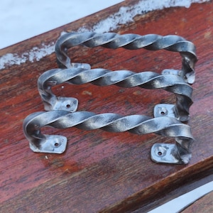 May include: Two handcrafted, twisted metal door handles. The handles are silver-toned with a textured, rope-like design. Each handle has a rectangular base with pre-drilled holes for mounting. The handles are displayed on a weathered, reddish-brown wooden surface.