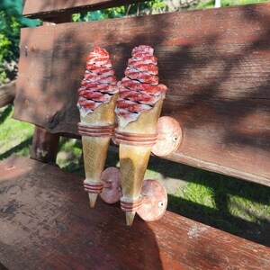 May include: Two decorative ice cream cone wall sconces. The cones are a light tan color with a waffle pattern. The ice cream is red and white, with a textured appearance. The sconces are mounted on a wooden bench.