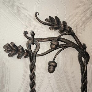 Peut inclure: Support d&eacute;coratif en m&eacute;tal fonc&eacute; orn&eacute; de feuilles de ch&ecirc;ne et de glands. Il pr&eacute;sente une base en colonne torsad&eacute;e et un sommet incurv&eacute; avec des d&eacute;tails de feuilles et de glands. Le m&eacute;tal a un aspect vieilli et rustique.