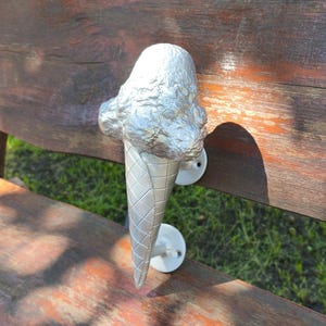 May include: A silver-colored metal door handle shaped like an ice cream cone with a textured scoop on top and a cross-hatched waffle cone base. The handle is attached to a surface by two circular white mounting plates with visible screw holes.