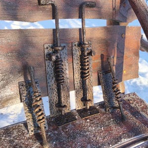 May include: Four rustic bronze metal latches. Each latch features a textured rectangular base, a spring, and a handle. The handles have a curved design. The latches are mounted on a wooden surface, suggesting a gate or door hardware.