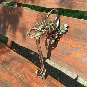 以下が含まれることがあります： 葉っぱのデザインが施されたサソリの形をした装飾的な金属製のドアノブ。ノブは暗い金属でできており、金色の仕上げが施されています。