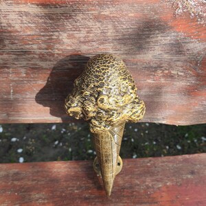 May include: A decorative ice cream cone door handle. The handle features a textured, golden-brown ice cream scoop atop a waffle cone. The handle is made of metal and is mounted on a wooden surface.