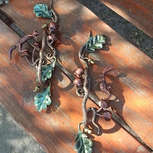 Puede incluir: Dos tiradores de puerta de metal decorativos con un tema de bosque. Cada tirador presenta una rama retorcida con hojas, setas y un animal pequeño. Un tirador tiene un zorro, el otro un conejo.