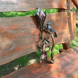 May include: A metal sculpture of a bird perched on a branch with two stylized flowers. The sculpture is painted in shades of brown, blue, and purple.