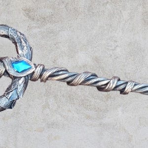 May include: A silver trident with a twisted handle and blue and green gemstones. The trident is a fantasy prop.