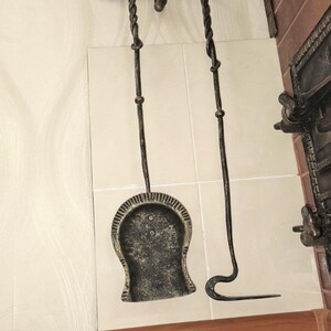Peut inclure: Un ensemble d'outils de chemin&eacute;e en fer forg&eacute;. L'ensemble comprend une pelle, un tisonnier et un dessus d&eacute;coratif avec des feuilles et des glands. Les outils sont gris fonc&eacute; et sont expos&eacute;s contre un mur carrel&eacute; blanc.
