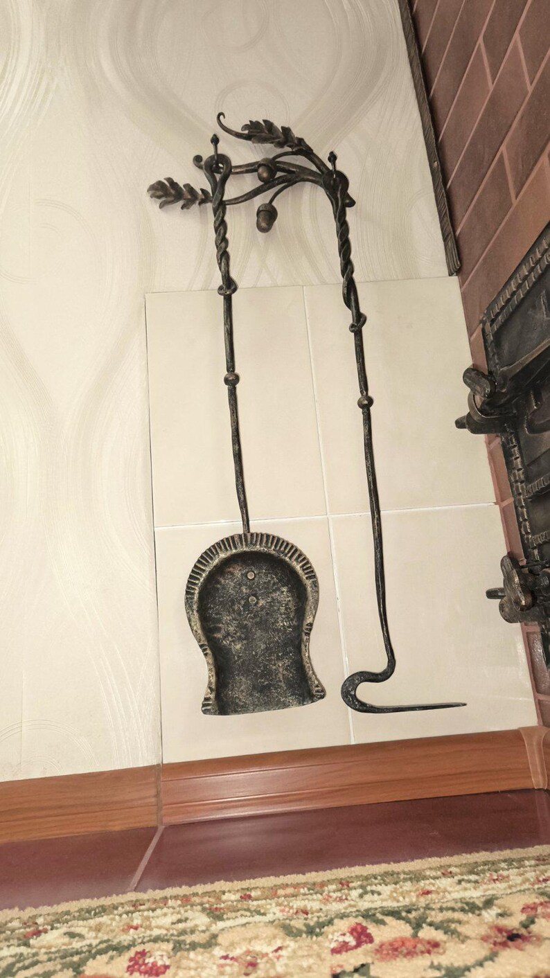 Peut inclure: Un ensemble d'outils de chemin&eacute;e en fer forg&eacute;. L'ensemble comprend une pelle, un tisonnier et un dessus d&eacute;coratif avec des feuilles et des glands. Les outils sont gris fonc&eacute; et sont expos&eacute;s contre un mur carrel&eacute; blanc.
