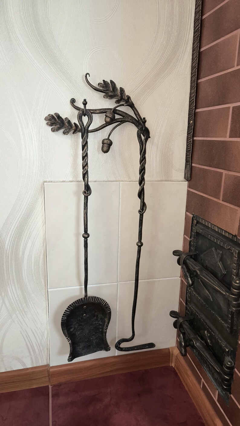 Peut inclure: Ensemble d'outils de chemin&eacute;e forg&eacute;s &agrave; la main. L'ensemble comprend une pelle et un tisonnier, tous deux avec des poign&eacute;es torsad&eacute;es et des accents d&eacute;coratifs de feuilles et de glands. Les outils sont en m&eacute;tal fonc&eacute; et mont&eacute;s sur un mur.
