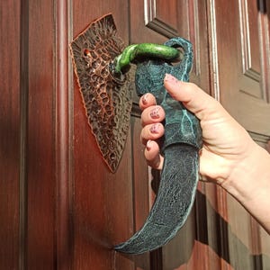 May include: A decorative door knocker with a dagger-shaped handle and a textured, diamond-shaped backplate. The handle is dark green and black, and the backplate is bronze. The knocker is mounted on a wooden door.