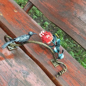 Puede incluir: Adorno de banco de metal con un pájaro posado en una rama con una flor roja y hojas verdes. El adorno está montado en un banco de madera con un acabado marrón rojizo. El pájaro tiene detalles plateados y negros.