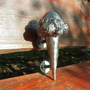 May include: A silver-colored ice cream cone door handle. The handle features a textured scoop of ice cream atop a cone with a diamond pattern. The handle is mounted on a wooden surface with two circular metal plates.