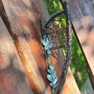 May include: A metal wall hook featuring a textured branch-like handle, a spiderweb design, and two decorative teal-colored oak leaves attached to a hammered rectangular base plate with a dark bronze finish and visible metallic highlights.