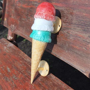 May include: A wall-mounted door handle shaped like an ice cream cone with three scoops in red, white, and green. The cone features a textured waffle pattern, and the handle is secured to the surface by two circular metal plates with screw holes.