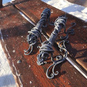 May include: Two ornate, dark gray metal handles with swirling, decorative elements. The handles are placed on a weathered, reddish-brown wooden surface, with a snowy background visible.