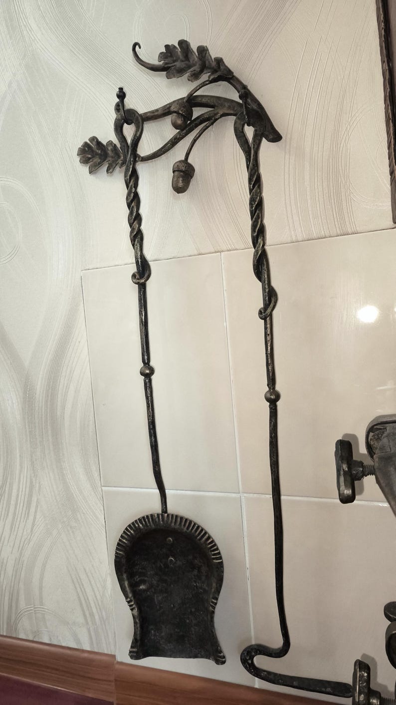 Peut inclure: Un ensemble d'outils de chemin&eacute;e en fer forg&eacute; avec un motif de feuilles de ch&ecirc;ne et de glands. L'ensemble comprend une pelle et un tisonnier avec des poign&eacute;es torsad&eacute;es et une finition sombre et vieillie. Les outils sont fix&eacute;s au mur.