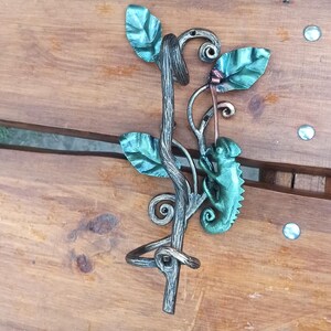 May include: A metal wall hook shaped like a vine with a chameleon climbing on it. The vine has green leaves and the chameleon is green with a brown stripe down its back.