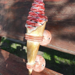 Op de afbeelding: Decoratieve wandlamp in de vorm van een ijshoorntje. De kegel is lichtbruin met een wafelpatroon. Het ijs is rood en wit. De lamp heeft twee ronde montageplaten. Het algehele ontwerp is grillig en leuk.