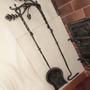 Peut inclure: Ensemble d'outils de chemin&eacute;e forg&eacute;s &agrave; la main. L'ensemble comprend une pelle, un tisonnier et un support d&eacute;coratif avec des accents de feuilles de ch&ecirc;ne et de glands. Les outils sont en m&eacute;tal fonc&eacute; et sont expos&eacute;s sur une surface carrel&eacute;e.