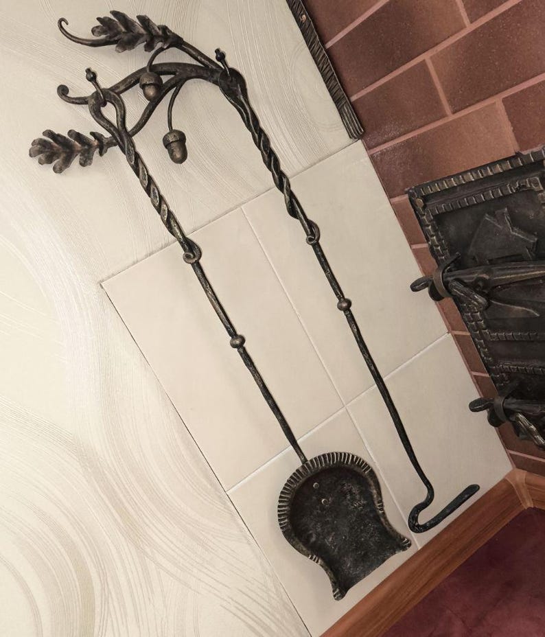 Peut inclure: Ensemble d'outils de chemin&eacute;e forg&eacute;s &agrave; la main. L'ensemble comprend une pelle, un tisonnier et un support d&eacute;coratif avec des accents de feuilles de ch&ecirc;ne et de glands. Les outils sont en m&eacute;tal fonc&eacute; et sont expos&eacute;s sur une surface carrel&eacute;e.