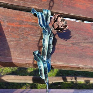 Puede incluir: Una escultura de metal de una flor con tallo y hojas. Las hojas son azules y verdes, y la flor es marrón. La escultura está colocada sobre un banco de madera.