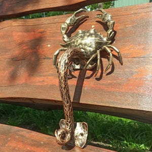 Puede incluir: Un tirador de puerta de bronce con forma de cangrejo con cola enroscada. El cangrejo está detallado con pinzas texturizadas y una concha texturizada.