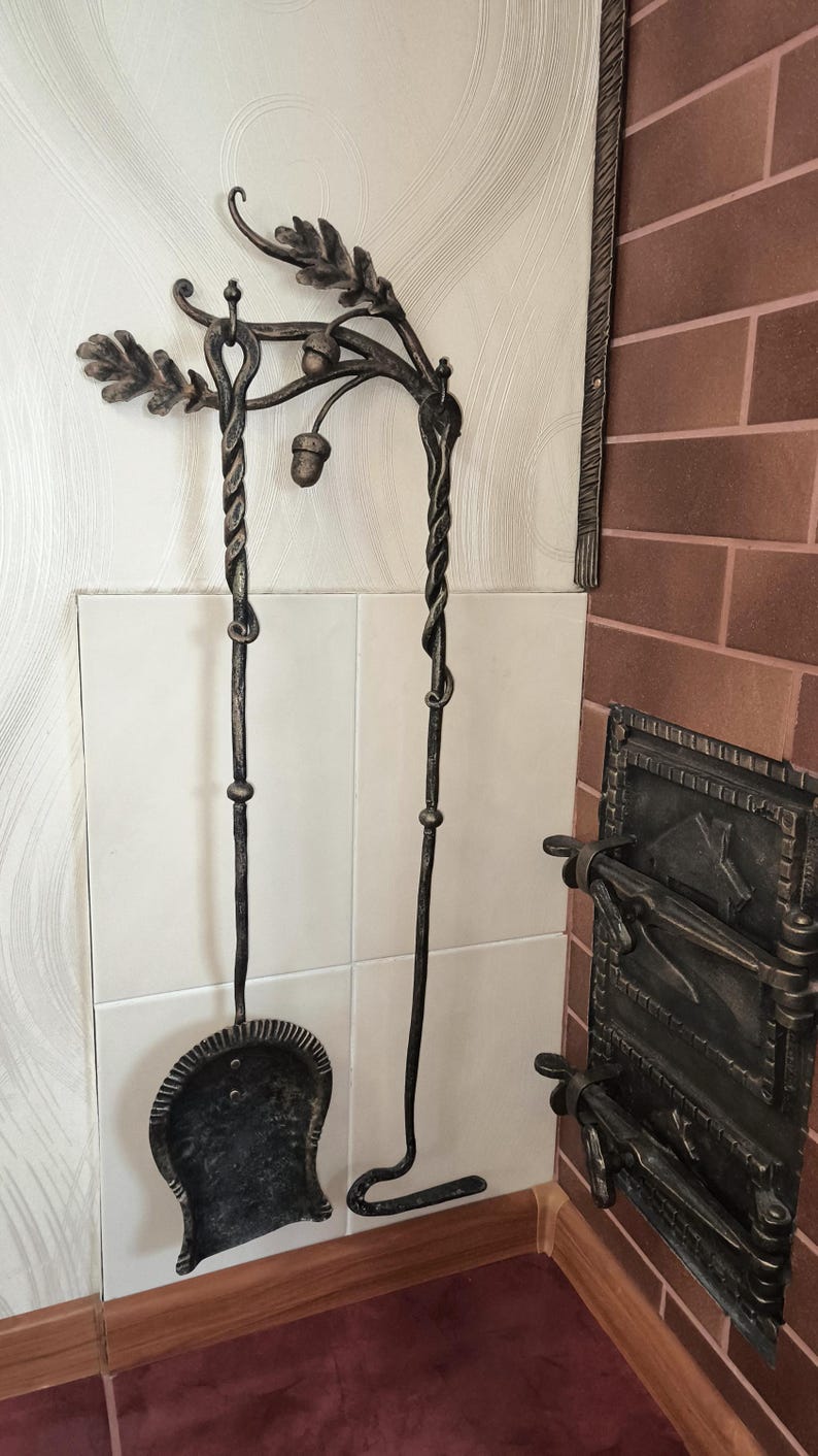 Peut inclure: Ensemble d'outils de chemin&eacute;e forg&eacute;s &agrave; la main. L'ensemble comprend une pelle et un tisonnier, tous deux avec des poign&eacute;es torsad&eacute;es et des d&eacute;corations de feuilles de ch&ecirc;ne et de glands. Les outils sont en m&eacute;tal fonc&eacute; et mont&eacute;s sur un mur carrel&eacute; blanc.