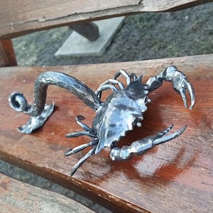 May include: A handcrafted metal crab sculpture with a textured finish. The crab has detailed claws, legs, and a shell. The sculpture is placed on a wooden surface, showcasing its intricate design and craftsmanship.
