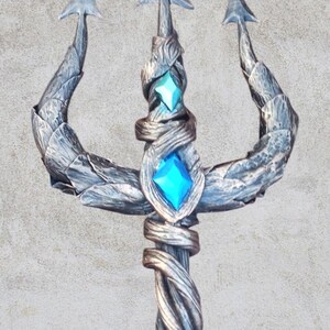 May include: A silver-colored trident with a twisted handle and two blue gemstones. The trident is made of metal and has a rustic, weathered look.