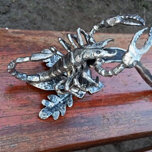 以下が含まれることがあります： 詳細な銀色の金属製のサソリの彫刻で、複雑なディテールが施されています。 サソリは葉の形をした台座の上に配置され、ハサミが伸びています。 彫刻は木製の表面に置かれています。