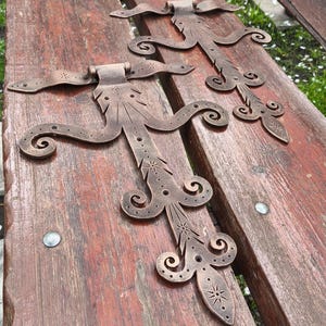 Puede incluir: Dos bisagras de metal de color bronce adornadas con diseños decorativos de estrellas y espirales. Las bisagras están dispuestas sobre una superficie de madera rojiza y envejecida. Las bisagras tienen una estética vintage y antigua.