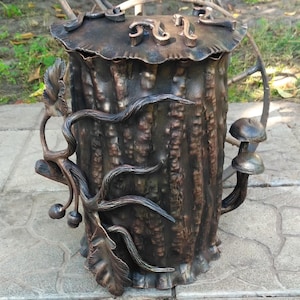 May include: A handcrafted metal bin with a tree trunk design. The bin features a lid with decorative scrollwork and is adorned with metal leaves, vines, and mushrooms. The color is a mix of brown and bronze.