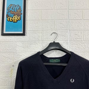 Może przedstawiać: Granatowy sweter z dekoltem w serek z małym białym logo na piersi, wiszący na czarnym wieszaku. W tle widoczny jest obraz w ramce z napisem "Life begins after coffee".