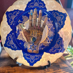 May include: Decorative blue plate with a hand illustration, featuring a Star of David and other symbols. The plate has a gold rim and ornate gold detailing. The hand has colorful markings on the fingers and is centered on the plate.