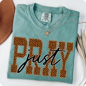 Könnte beinhalten: Ein türkisfarbenes Comfort Colors T-Shirt mit den Worten "PRAY just" in einem Leopardenmuster. Eine zarte Silberkette mit einem kleinen Anhänger liegt über dem Shirt. Das Shirt ist auf einer weißen Oberfläche gefaltet.