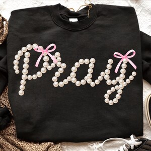 Könnte beinhalten: Schwarzes Sweatshirt mit dem Wort "Pray", das aus Kunstperlen gebildet wird. Die Buchstaben sind mit rosa Schleifen akzentuiert. Das Sweatshirt ist ein Rundhals-Modell.