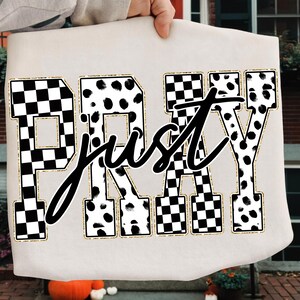 Könnte beinhalten: Cremefarbener Stoff mit den Worten "Just Pray" in schwarzer Schrift über Blockbuchstaben. Die Blockbuchstaben sind goldfarben umrandet und mit einem schwarz-weißen Schachbrettmuster oder einem schwarz-weißen Dalmatiner-Punktmuster gefüllt.