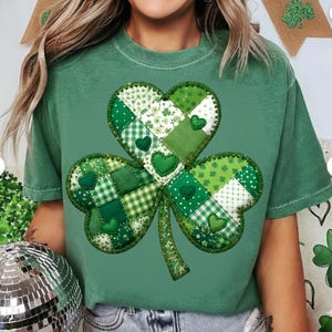 May include: Green t-shirt featuring a large shamrock design. The shamrock is made of various green patterned fabrics, including polka dots, floral prints, and gingham. Small green heart accents are scattered throughout the design. The shirt is a solid green color.