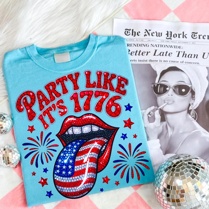 May include: A light blue t-shirt with the text "PARTY LIKE IT'S 1776" in red, featuring a red lip design with a tongue in the American flag pattern. The shirt has red and blue star and firework accents.