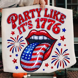 May include: A cream-colored tote bag featuring the text "PARTY LIKE IT'S 1776" in red, embellished with rhinestones. The design includes a red-lipped mouth with a tongue styled as the American flag, surrounded by red, white, and blue stars and fireworks.