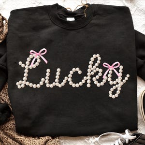 以下が含まれることがあります： 黒色のスウェットシャツ。フェイクパールで「Lucky」の文字が描かれています。文字には小さなピンクのリボンが2つ付いています。クルーネックのスタイルで、白い表面に表示されています。