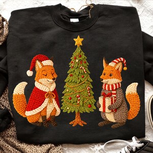 May include: Black sweatshirt with an embroidered Christmas design. Two foxes are featured, one in a Santa hat and cape, the other holding a gift. A decorated Christmas tree is between them, with a gold star on top.