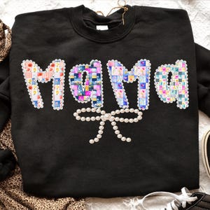 May include: Black sweatshirt with the word "MAMA" in colorful patchwork letters. A pearl bow is centered below the letters. The letters are outlined with a scalloped edge.