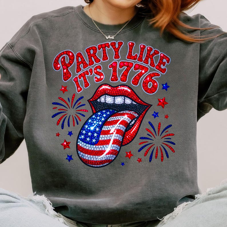 May include: A dark gray sweatshirt with the text "PARTY LIKE IT'S 1776" in red, white, and blue. The design features a pair of red lips with a tongue in the American flag design, surrounded by fireworks and stars.