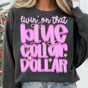 Może przedstawiać: Ciemnoszara bluza z różowym napisem "livin' on that blue collar dollar", obramowanym ciemniejszym różem. Tekst ozdobiony jest perłami i gwiazdkami. Bluza ma swobodny styl.
