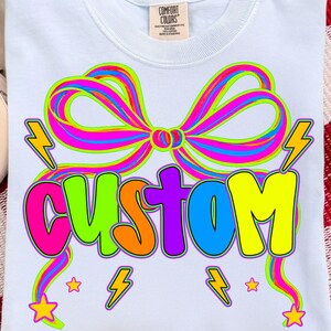 Op de afbeelding: Een lichtblauw t-shirt met een grote, veelkleurige afbeelding van een strik en het woord CUSTOM in dikke, levendige letters. Het ontwerp bevat neonkleuren zoals roze, groen, oranje, paars, blauw en geel, aangevuld met bliksemschichten en sterren.