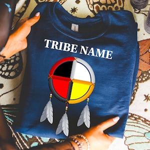 Puede incluir: Sudadera azul marino con el texto blanco "TRIBE NAME". Presenta un diseño de atrapasueños colorido con segmentos rojos, negros, blancos y amarillos, y plumas grises. Una prenda cómoda y elegante.