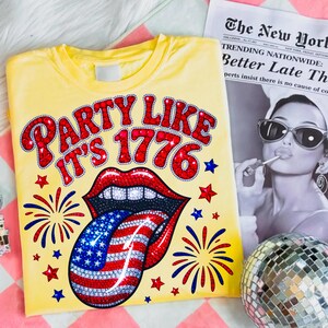 May include: A yellow t-shirt with the phrase "PARTY LIKE IT'S 1776" in red, rhinestone-studded letters. The design includes a mouth with a tongue styled as the American flag, surrounded by stars and fireworks.