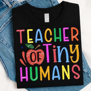 Puede incluir: Camiseta negra con el texto multicolor y lúdico "TEACHER OF TINY HUMANS". Un dibujo de una manzana está en el centro. La camiseta se muestra sobre un fondo de mezclilla.