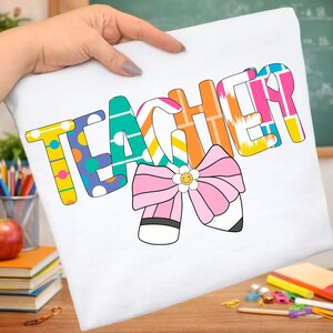 Pu&ograve; includere: T-shirt bianca con la scritta "TEACHER" in lettere colorate e decorate. Sotto la parola c'&egrave; un fiocco rosa con un centro a margherita e un disegno di matita. La maglietta &egrave; tenuta in mano.